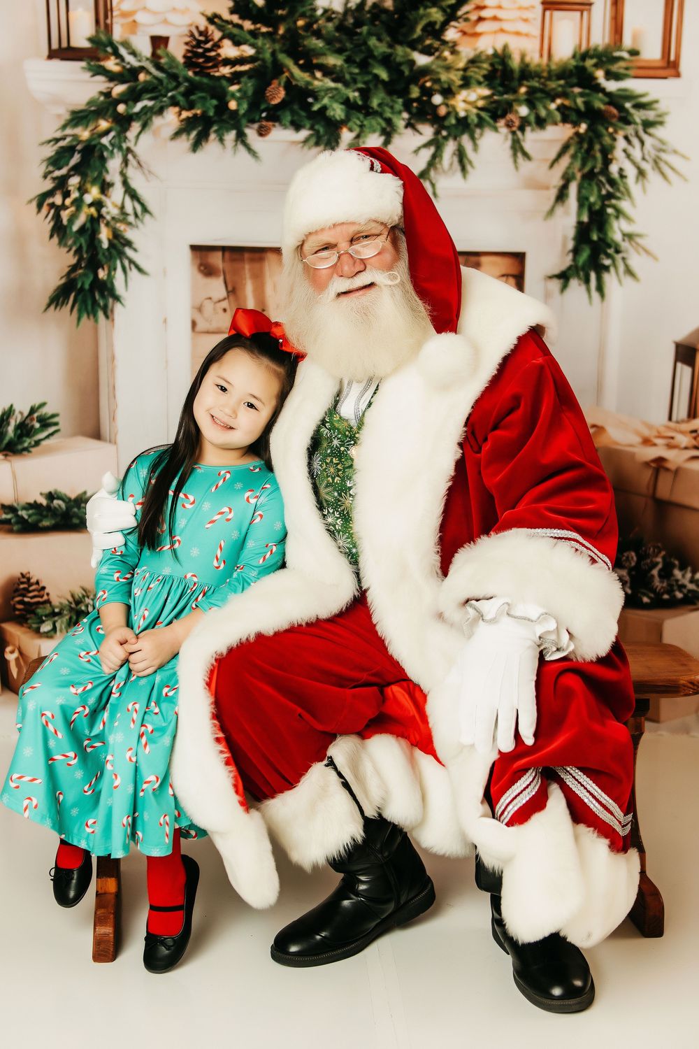 A child in a mint green dress poses with a jolly holiday figure in classic red and white costume by decorated greenery.
