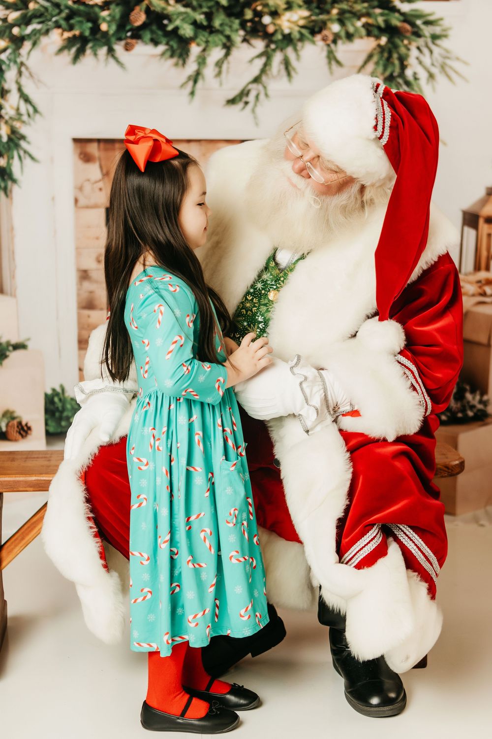 A child in a turquoise dress interacts with someone in a red and white holiday costume in a festive indoor setting.