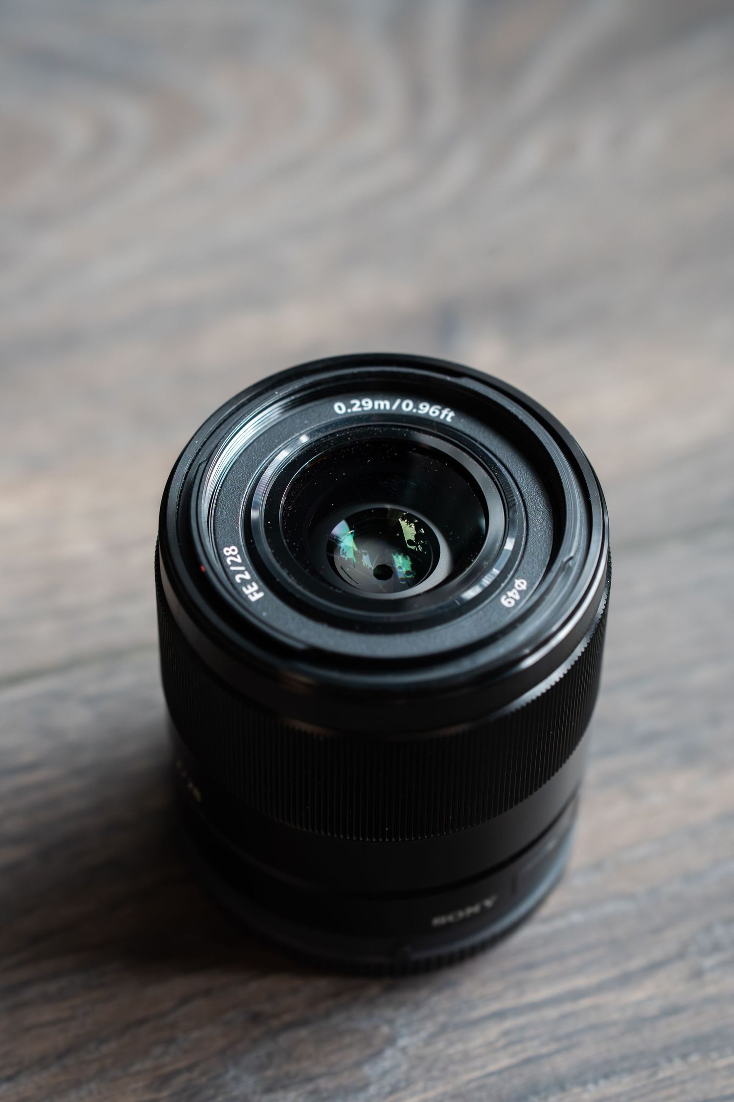 A black Sony camera lens with glass elements visible sits on a wooden surface in dramatic lighting.