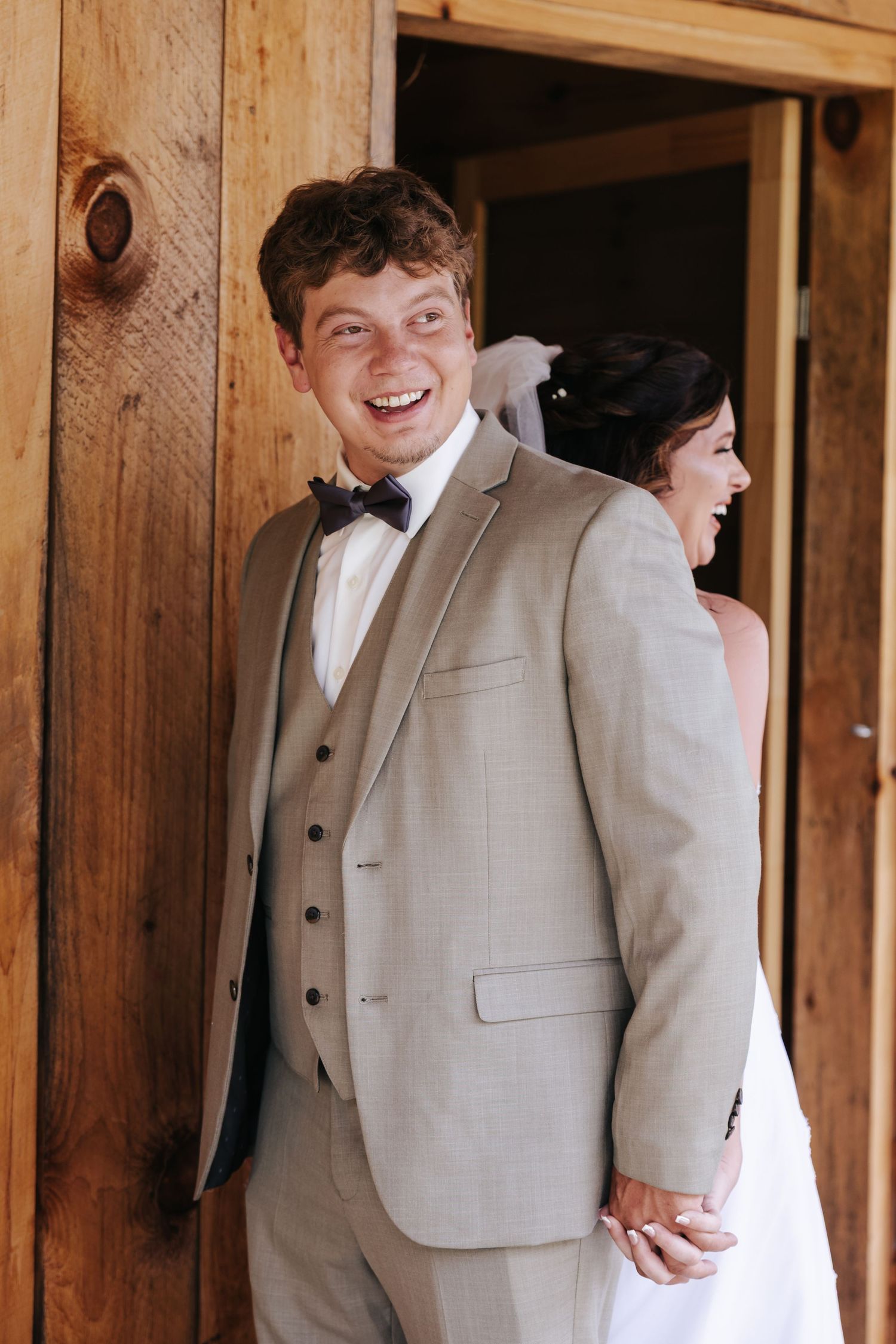 A group of wedding party members in light beige suits stand together and laugh in a rustic wooden setting.