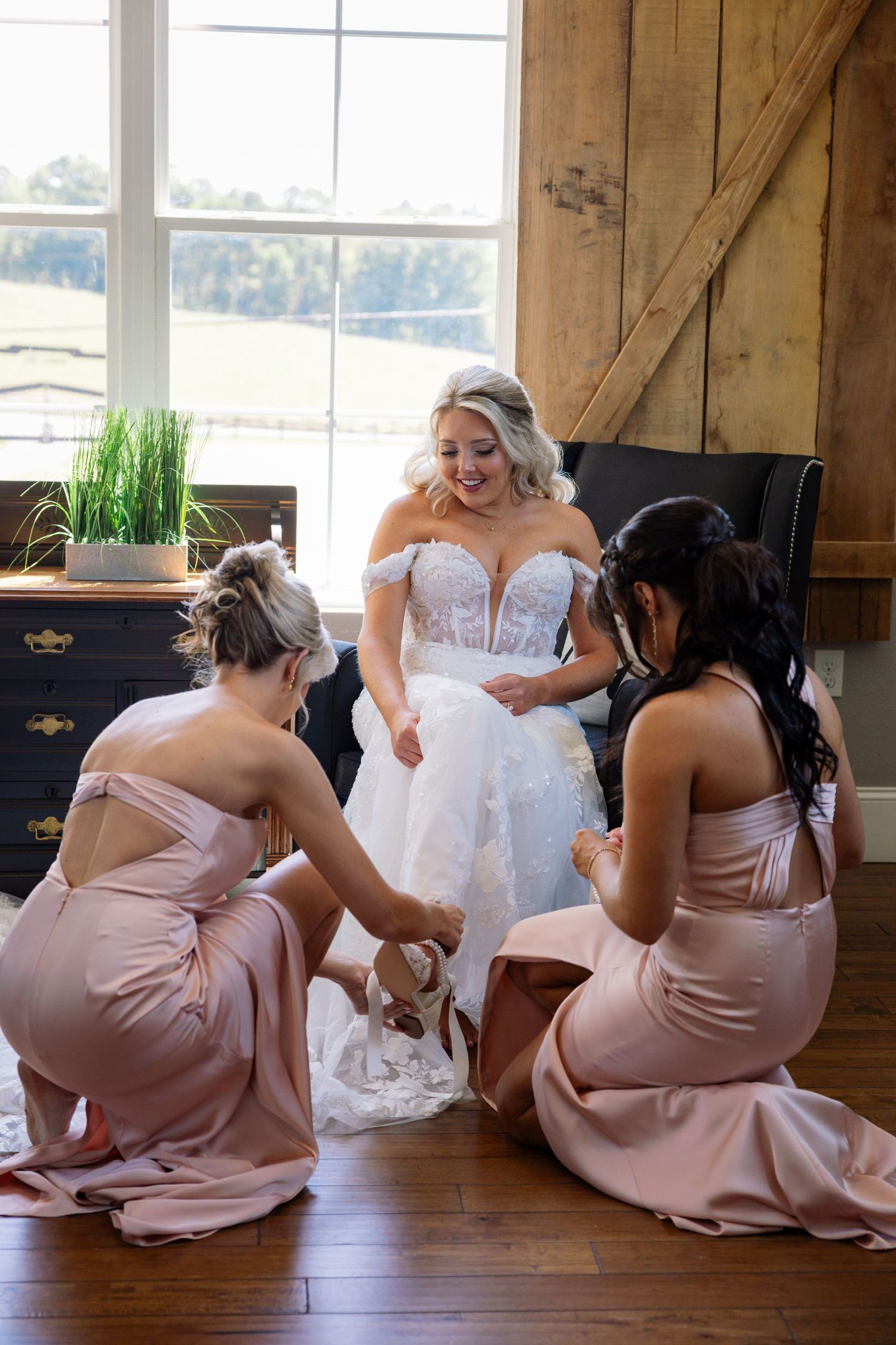 Bridesmaids in blush pink dresses sit on floor helping bride in white gown during wedding preparations.