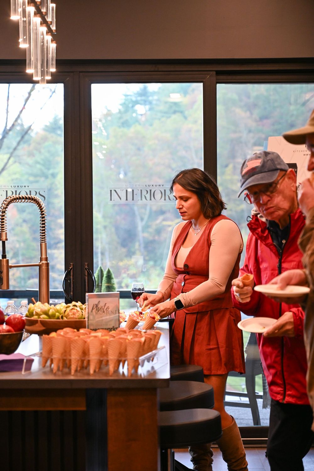 People browse a cozy interior space with pendant lighting and large windows overlooking nature.