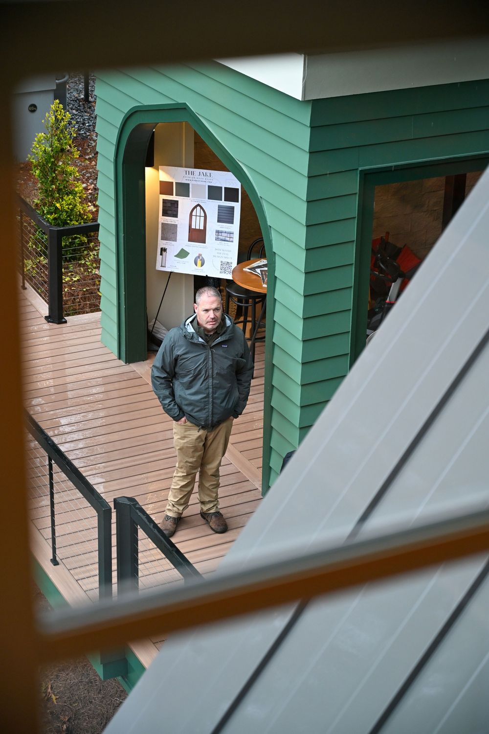 Someone stands on a wooden deck outside a green building entrance with a notice board visible.
