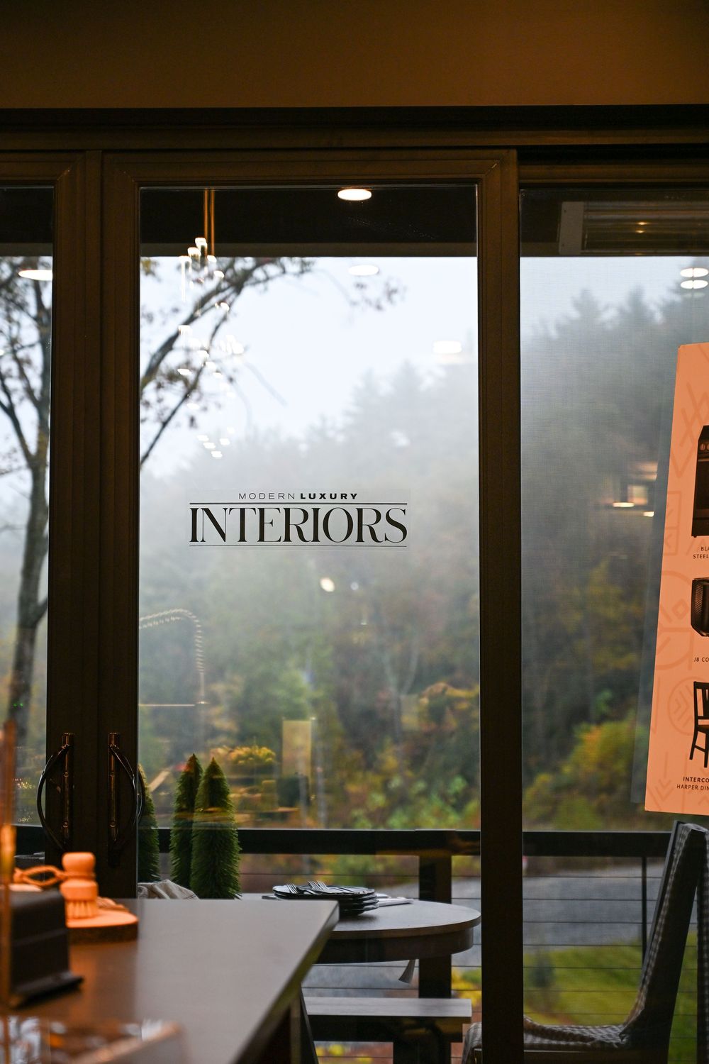 Interior view through large windows showing foggy forest landscape and signage for 'Interiors' in modern retail space.