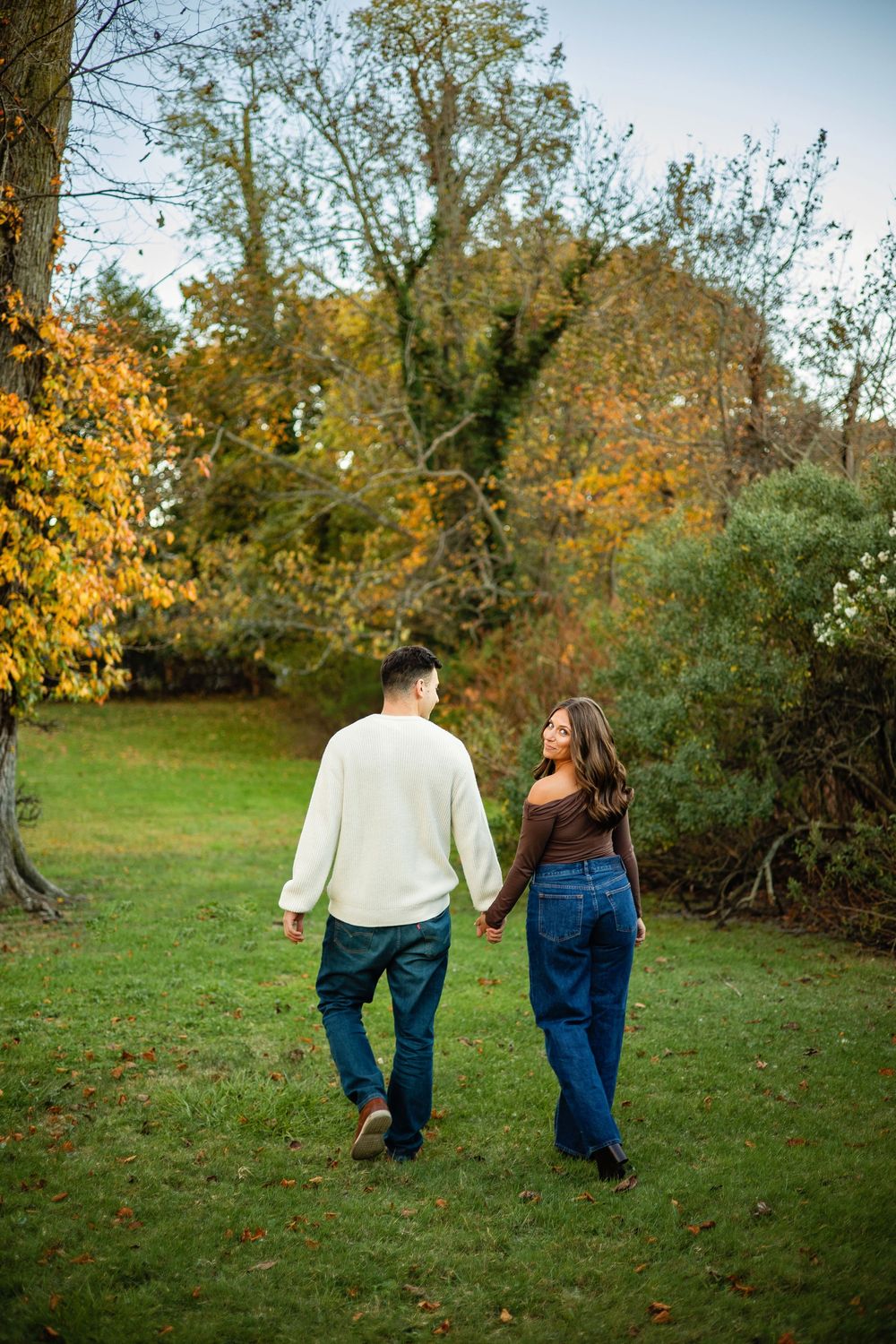 A couple in matching denim walks hand in hand through a scenic autumn park with golden foliage.