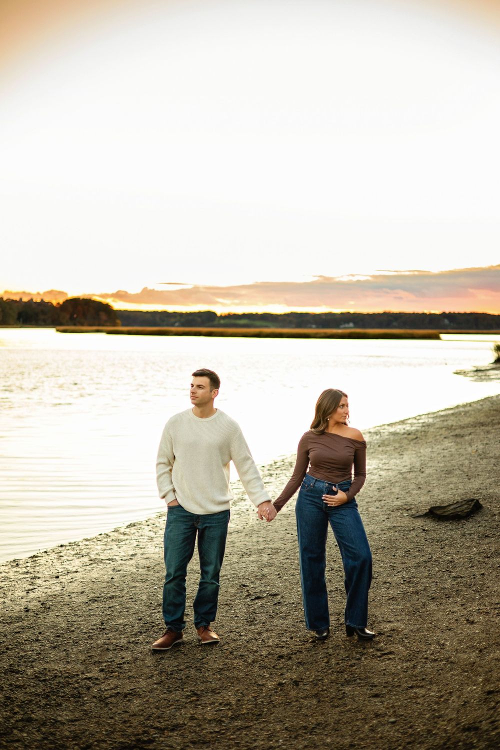 A couple holds hands while walking along a scenic beach shoreline at sunset with water and sky in warm golden light.