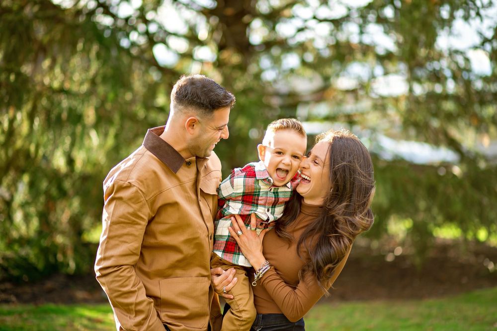 A couple in autumn attire shares tender moments in a park setting with sunlit trees in the background.