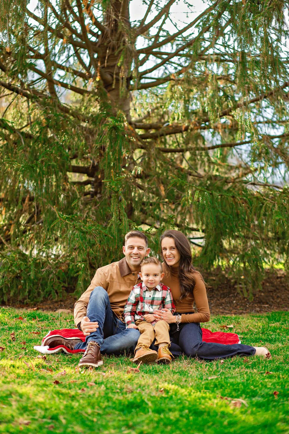 A family relaxes together on a red blanket under evergreen trees in a grassy outdoor setting.