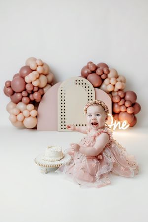 Baby girl in pink dress celebrates first birthday with cake and balloon arch backdrop.