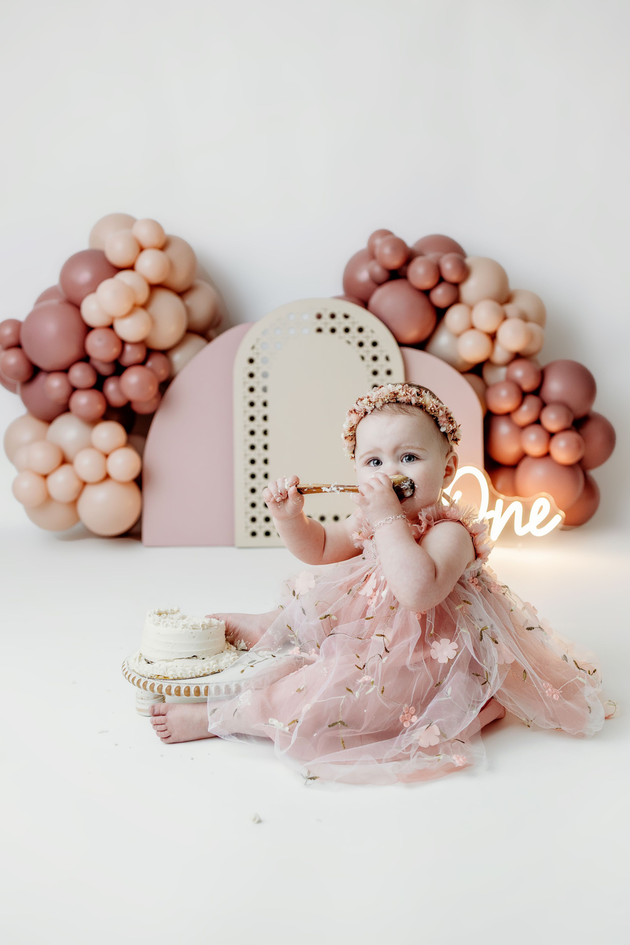 Baby girl in pink tulle dress celebrates first birthday smashing cake surrounded by mauve balloon garland and neon One sign.