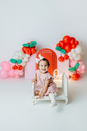 Baby girl in floral dress sits on white chair at cherry-themed first birthday party with balloon decorations.