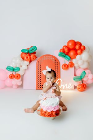 Baby girl in floral romper enjoys first birthday cake smash with cherry balloon decorations and neon “One” sign.
