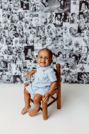 Smiling baby in blue romper sits on wooden chair before black and white photo collage backdrop celebrating first birthday.