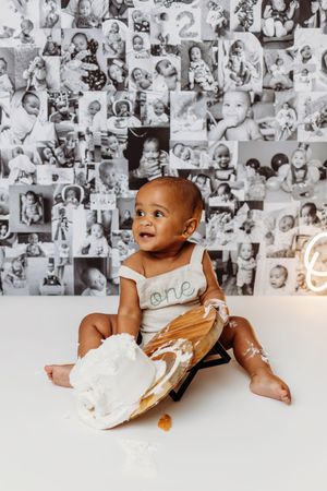 Baby's first birthday cake smash photo with smashed cake on white floor, wearing "one" onesie, black and white photos on wall.