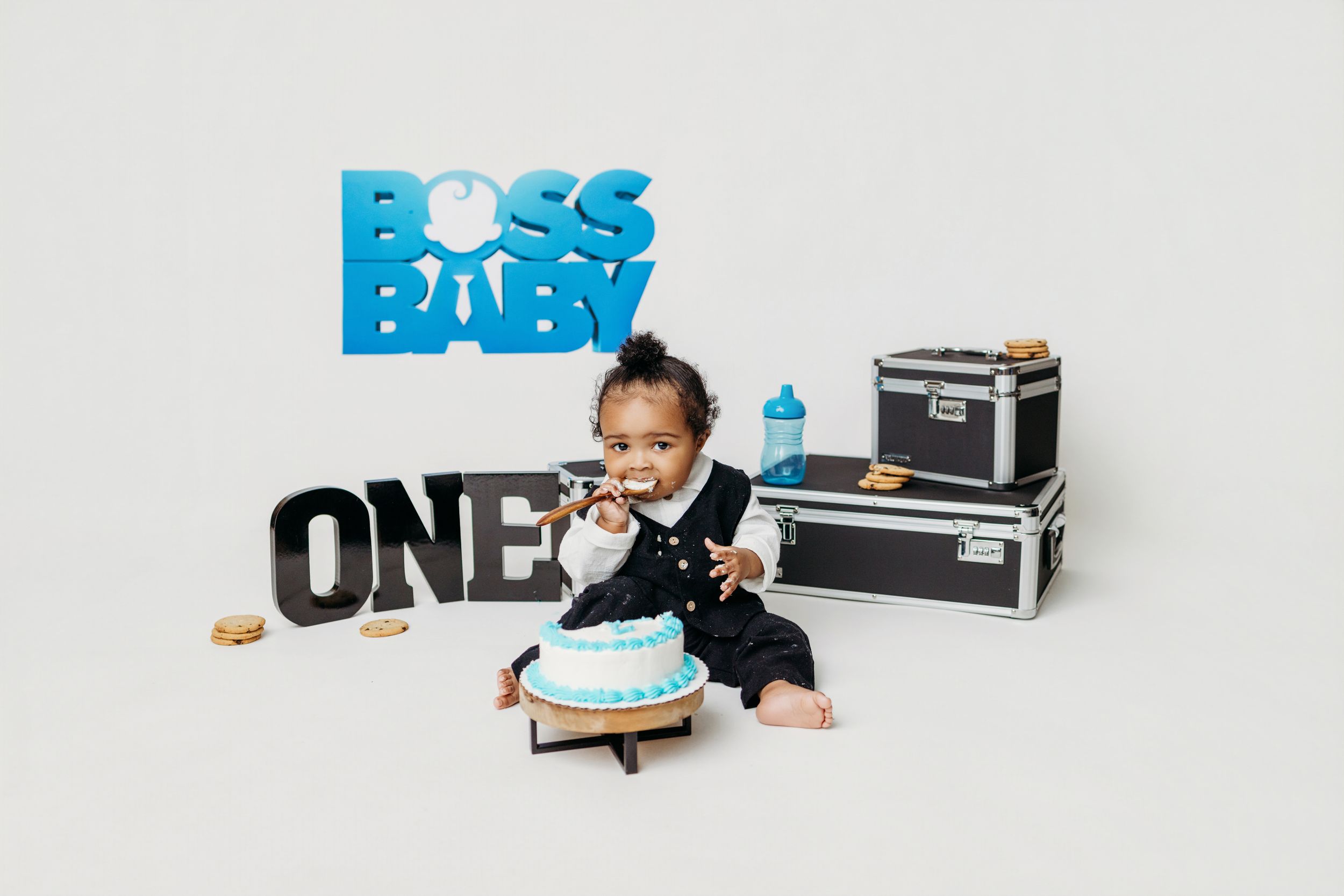 Baby in suit celebrates first birthday with cake smash, Boss Baby themed backdrop with blue letters and ONE sign.
