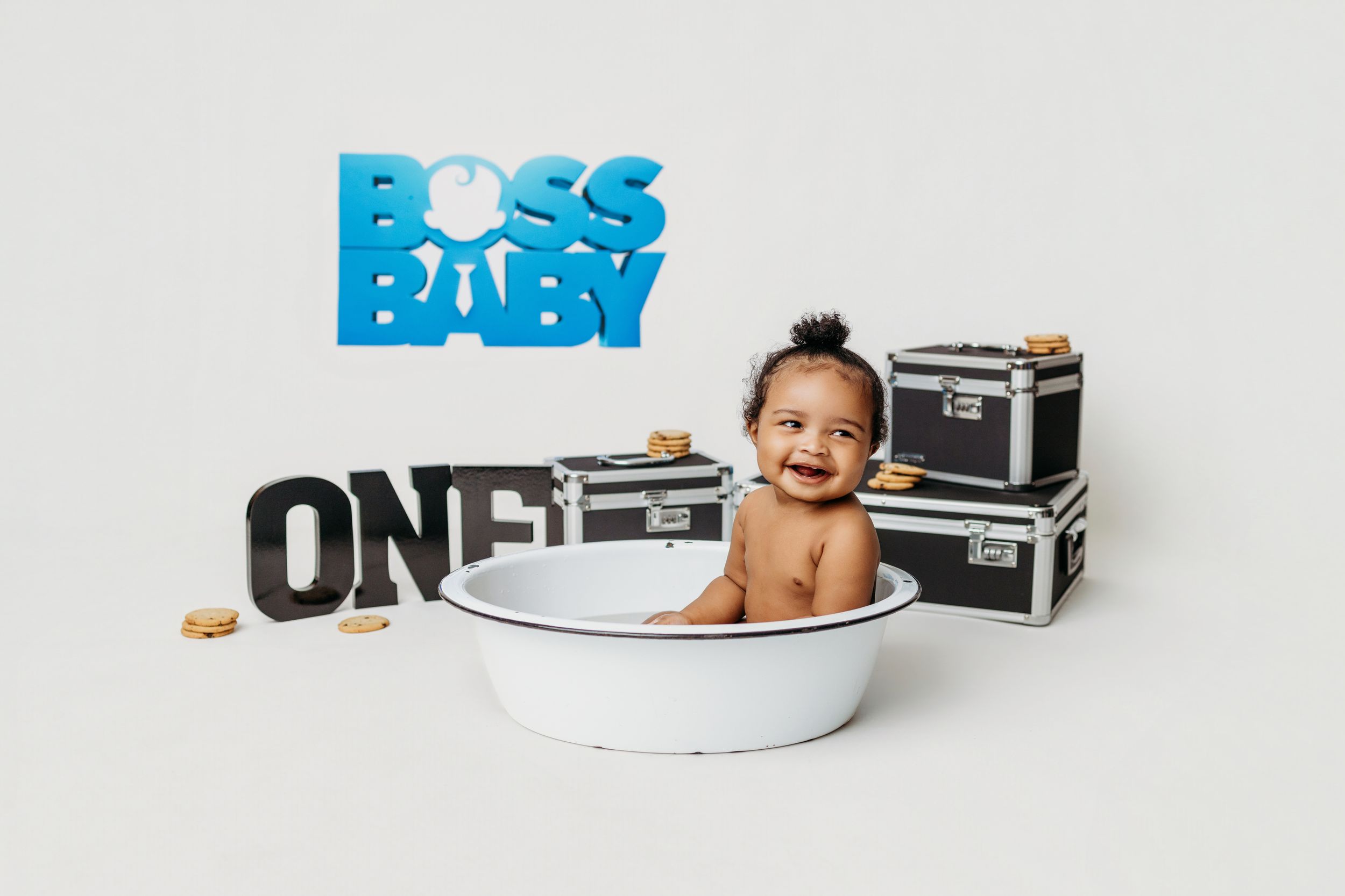 Smiling baby sitting in white tub with Boss Baby and ONE decorations on white background for first birthday photo.