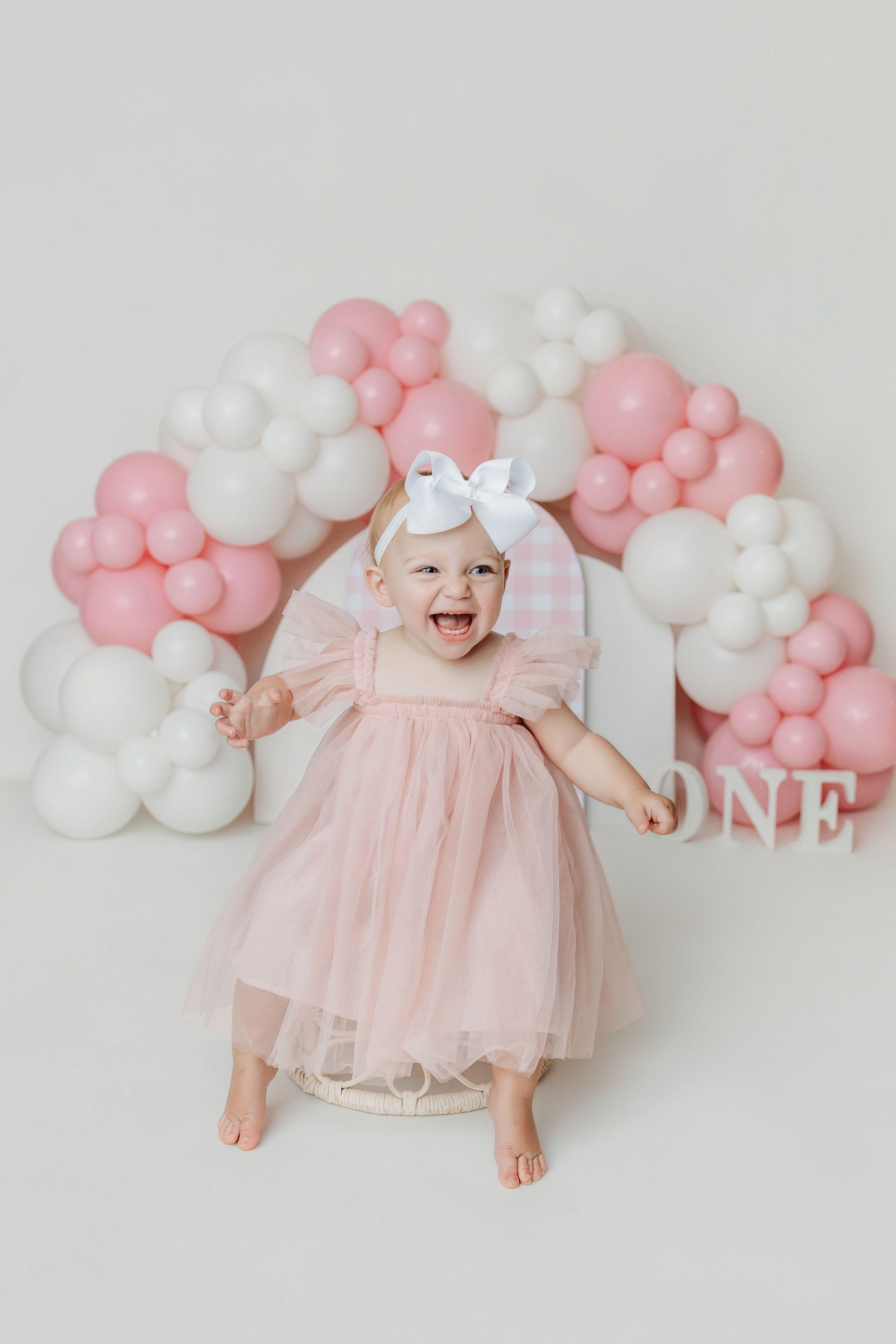 Happy baby girl in pink tulle dress celebrates first birthday with balloon arch backdrop.