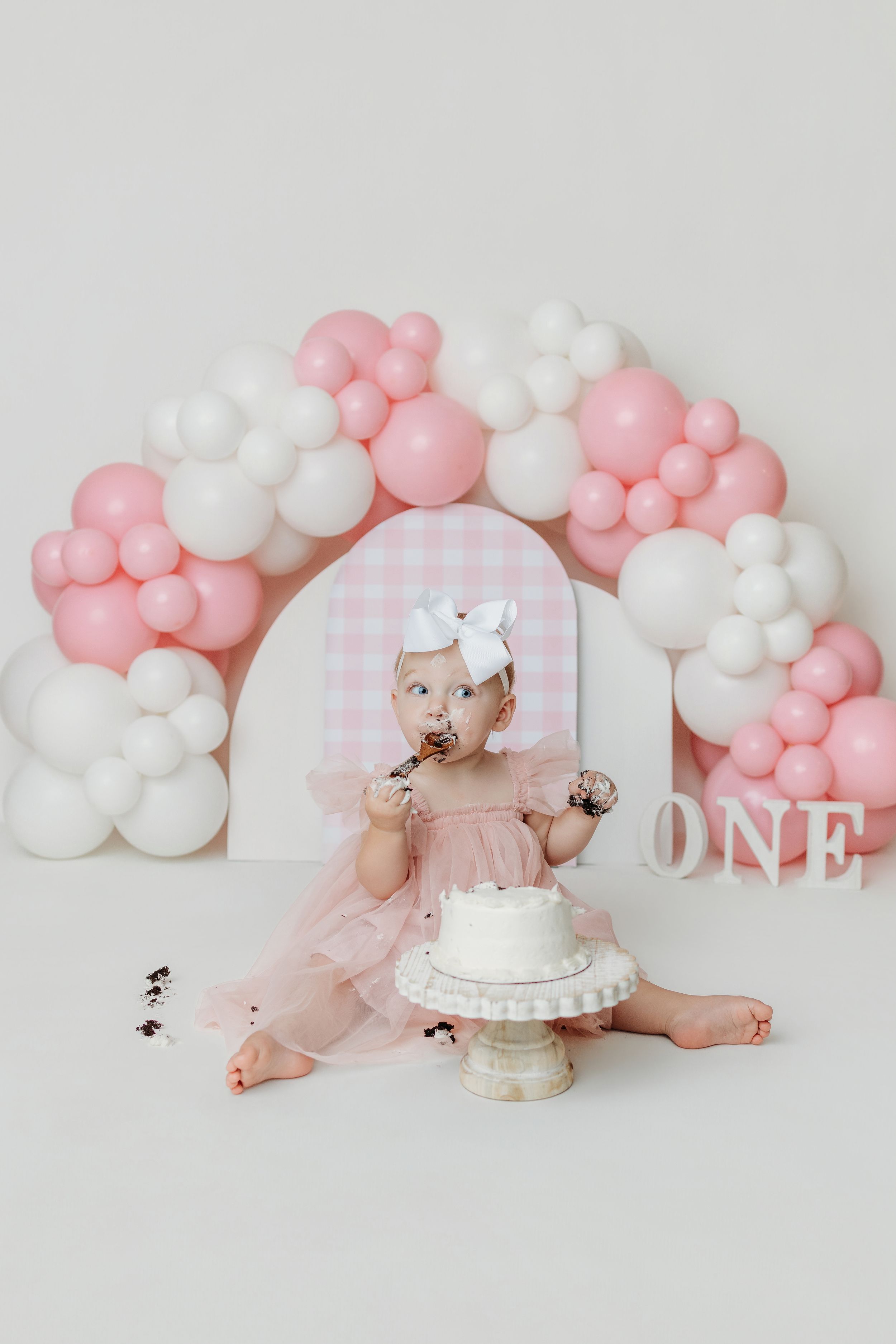 Baby girl in pink dress smashes first birthday cake surrounded by pink and white balloon arch with ONE sign.