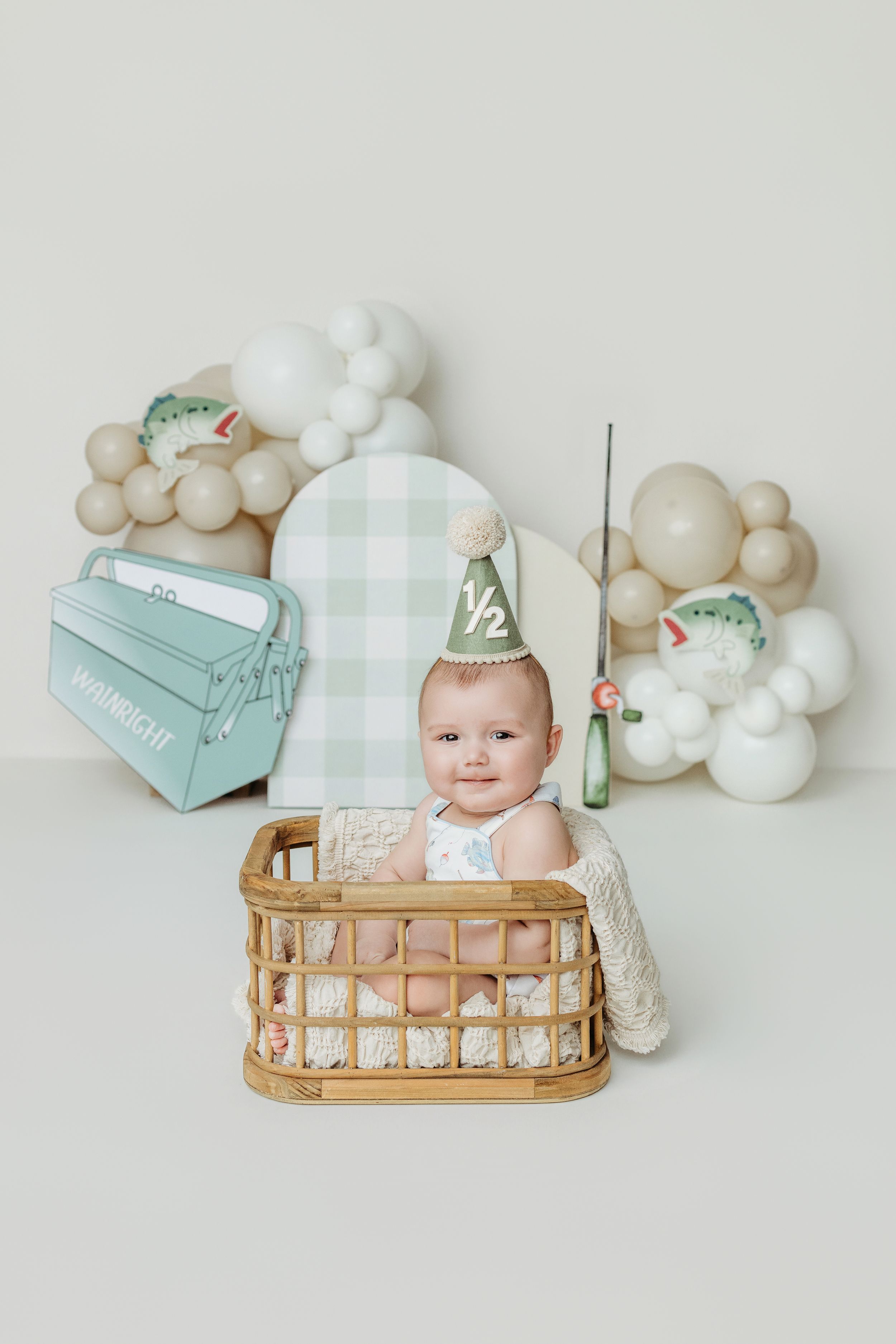 Adorable baby in wicker basket wearing half birthday party hat, surrounded by balloons and festive decorations.