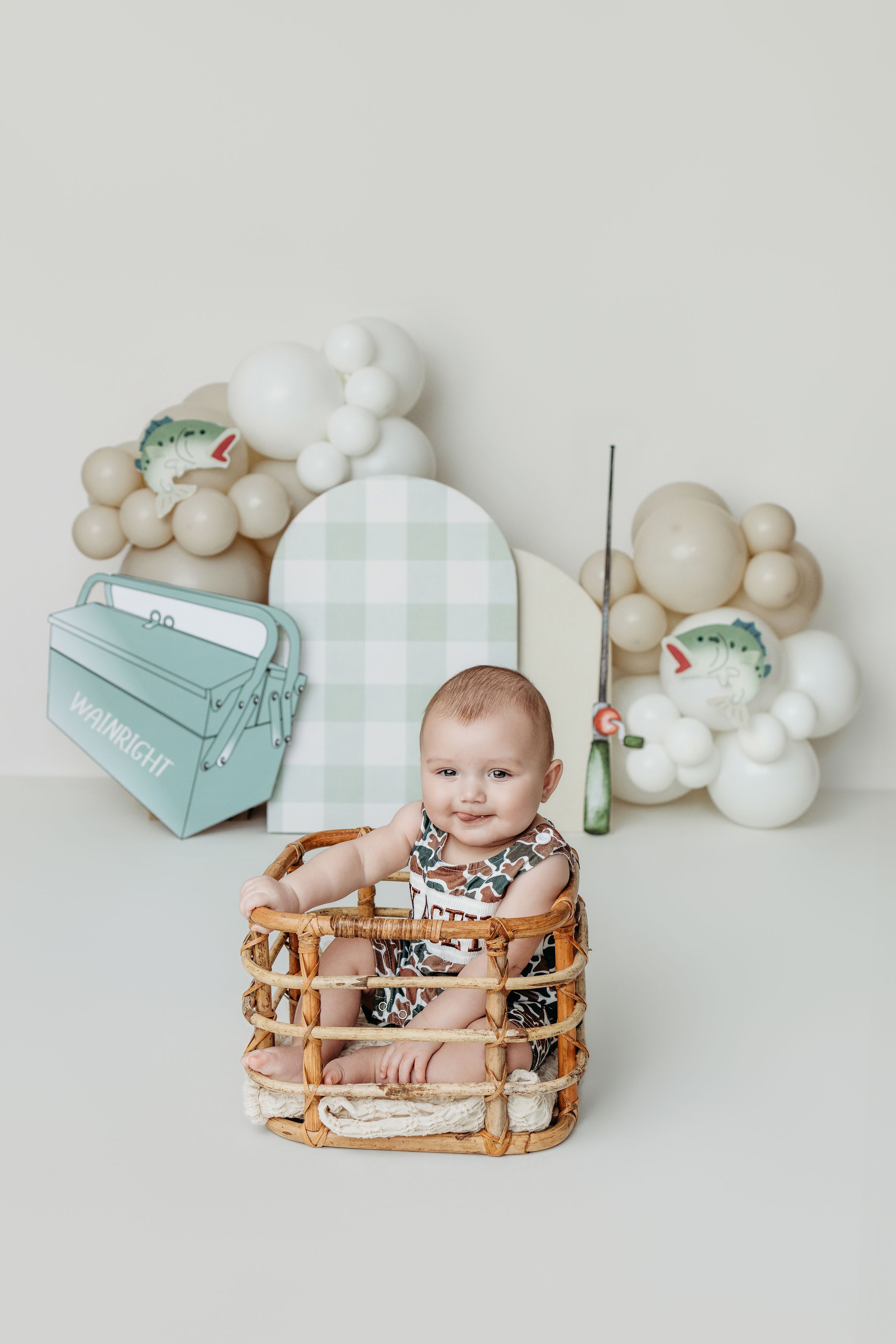 Smiling baby sitting in a wicker basket with balloon garland and fishing themed props on white backdrop.
