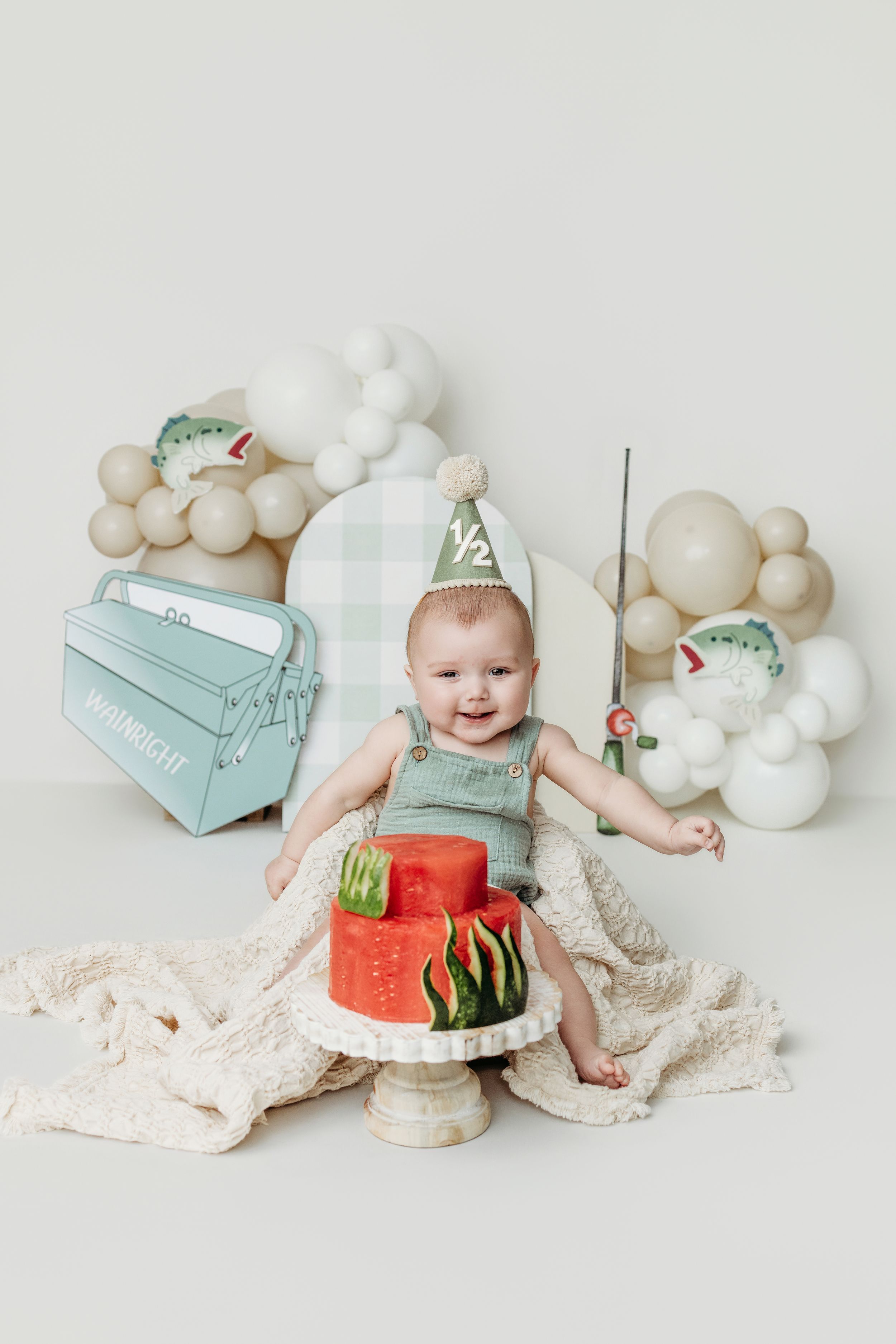Baby in fishing outfit celebrates half birthday with watermelon cake, balloons, and tackle box decor.