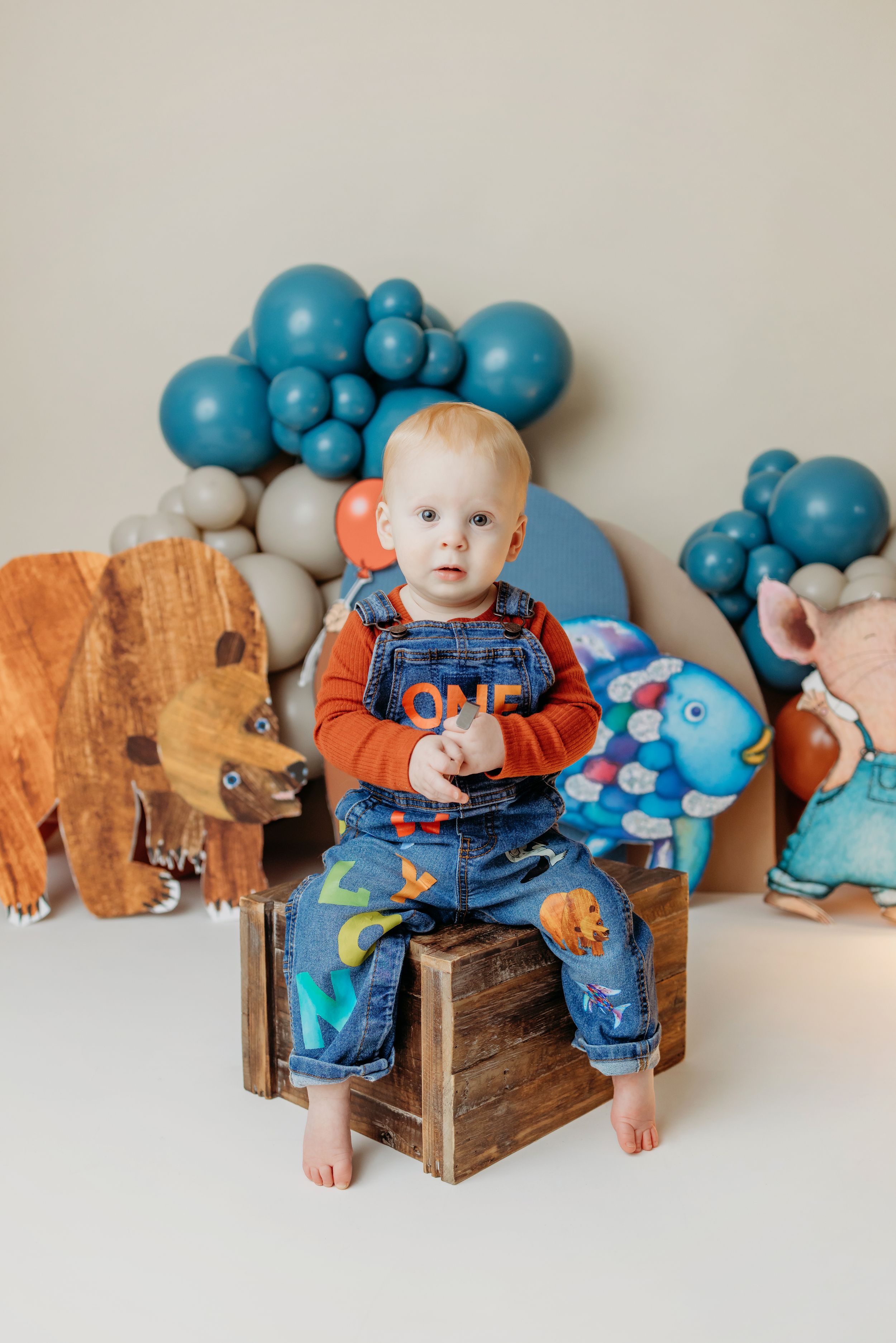 Toddler boy in denim overalls sitting on wooden crate at first birthday party with blue balloons and animal decorations.