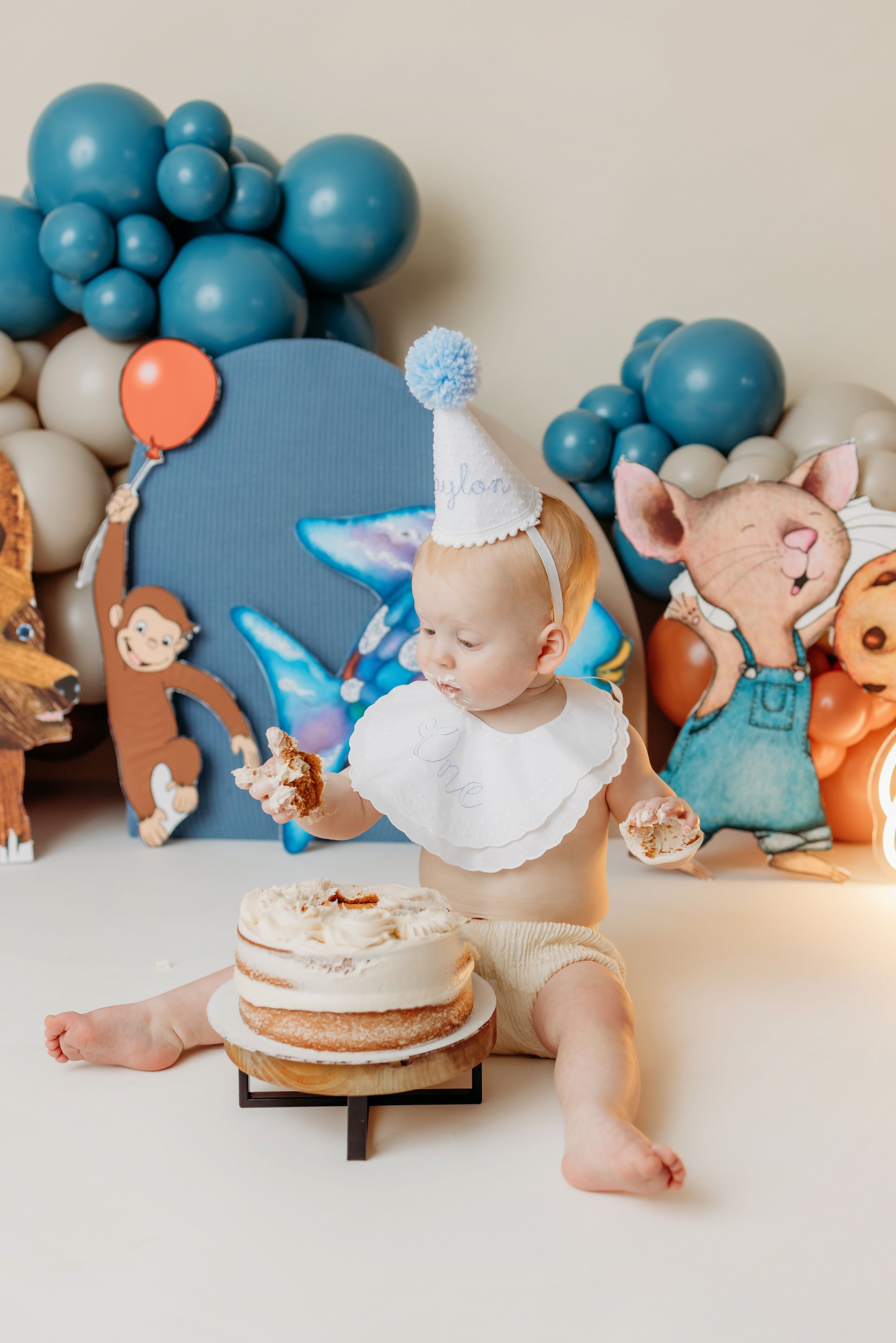 Baby in party hat sits before smash cake surrounded by animal cutouts and blue balloon garland decor.