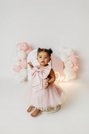 Toddler girl in pink tulle dress with bow at first birthday party with balloon arch and neon One sign.