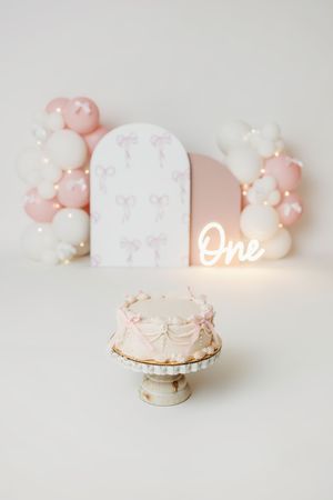 Pink and white first birthday cake smash setup with balloon arch, bow decorations, and glowing "One" neon sign.
