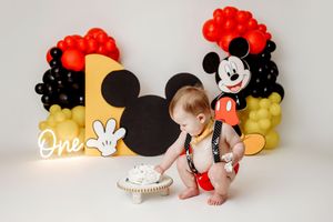 Baby's first birthday cake smash with Mickey Mouse theme, balloons, and decorations spelling One.