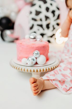 Pink birthday cake with silver ornaments on a cake stand, baby feet visible, first birthday smash cake celebration.