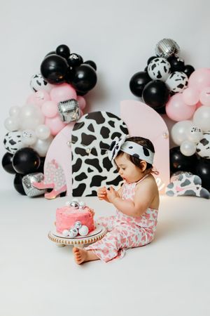 Baby girl in cow print outfit celebrates first birthday with pink cake smash, surrounded by black and pink balloon garland.
