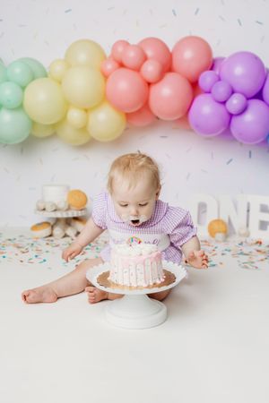 Baby girl celebrating first birthday with white cake, rainbow balloon arch, and colorful confetti decorations.