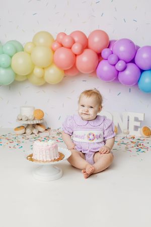 Baby girl in purple outfit sits beside a pink smash cake, surrounded by rainbow balloons and confetti for her first birthday.
