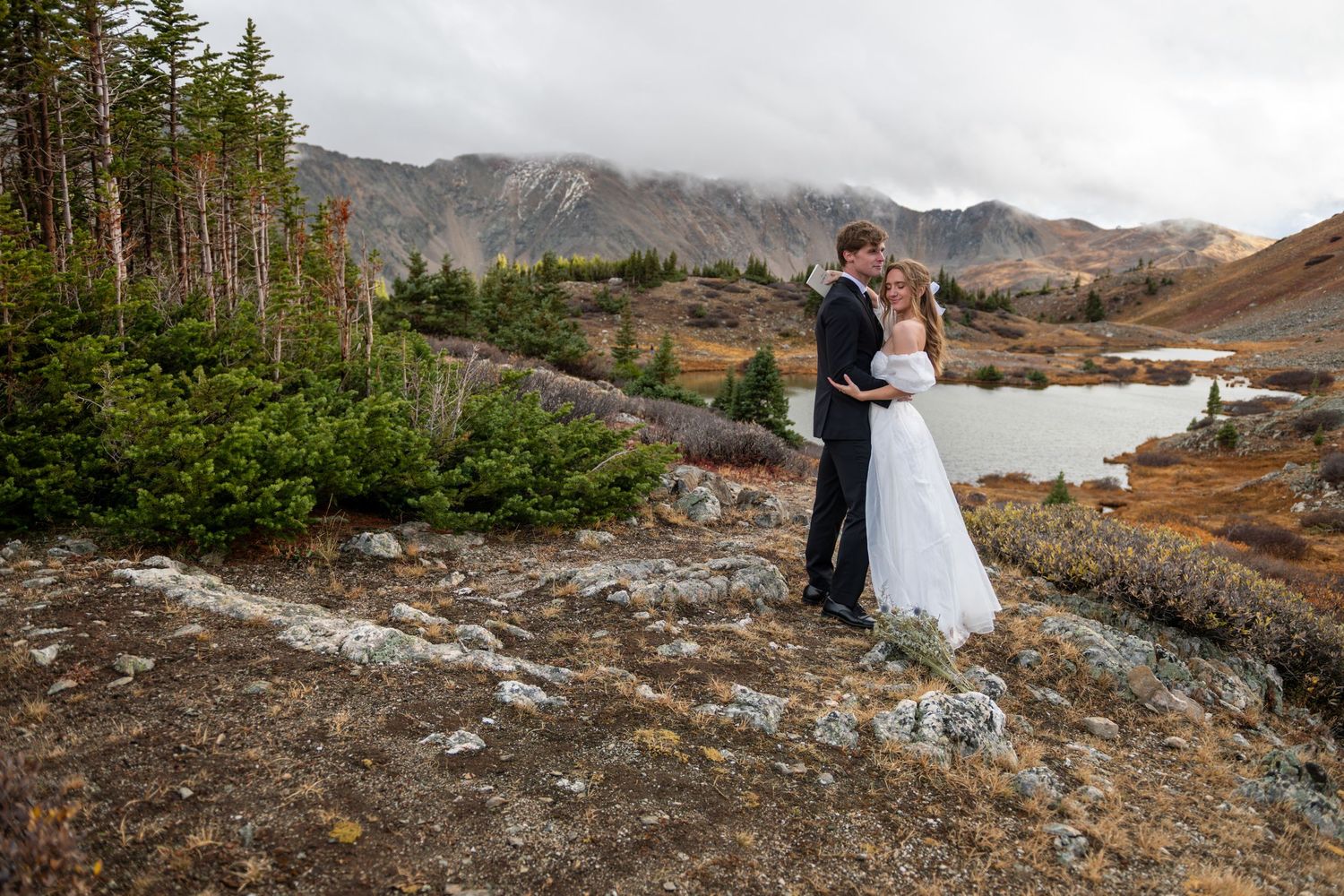 A bride and groom stand hugging on a scenic autumn mountain trail with rugged mountains behind them in fog.