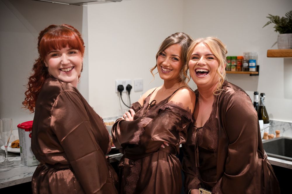 Three people wearing matching brown robes laugh together in a kitchen setting.