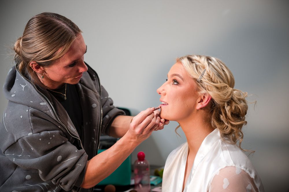 Professional makeup artist applies finishing touches to a bride's elegant wedding day look in a soft-lit setting.