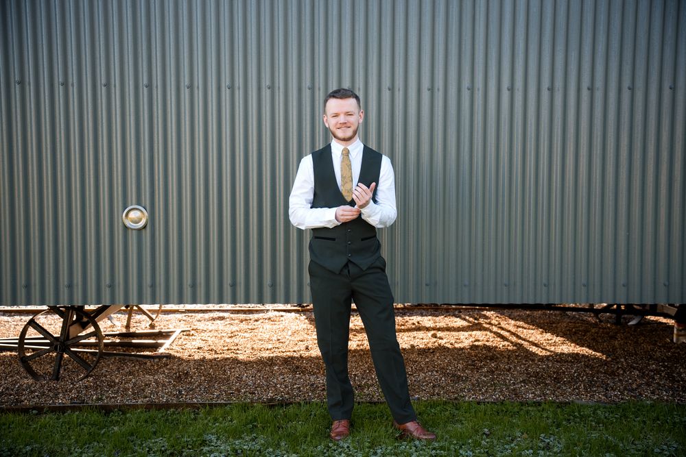 A person in formal attire stands in front of a corrugated metal wall wearing a vest and tie.