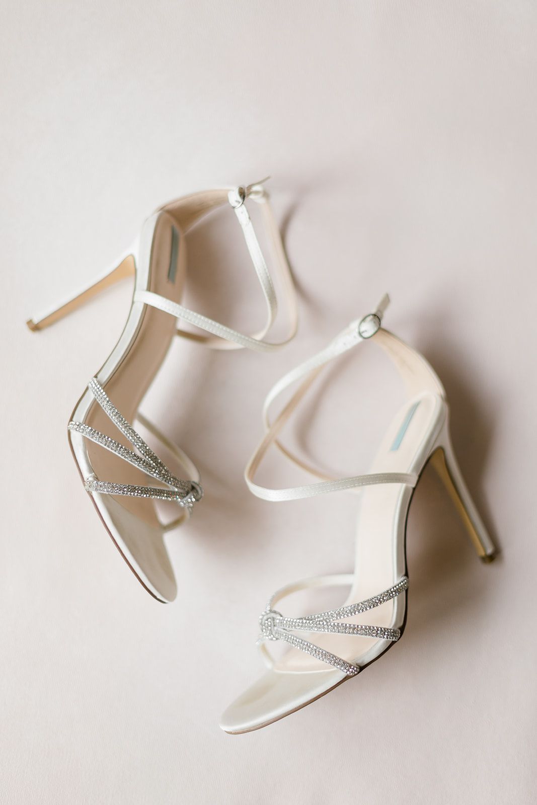 Elegant white satin bridal heels with crystal embellishments and ankle straps displayed on light background.