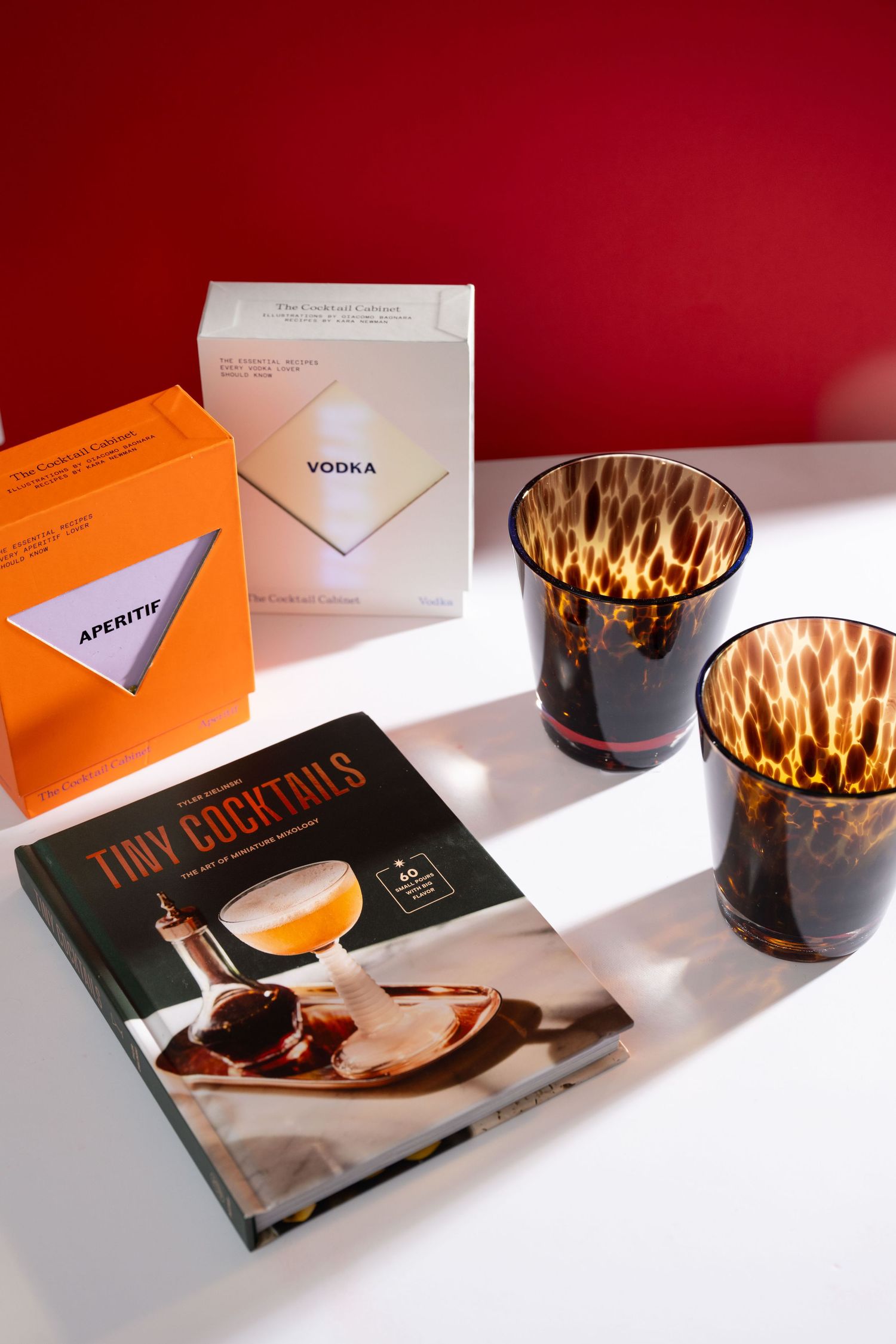 Stylish cocktail menu and patterned glass tumblers arranged on white surface against red wall with dramatic lighting.