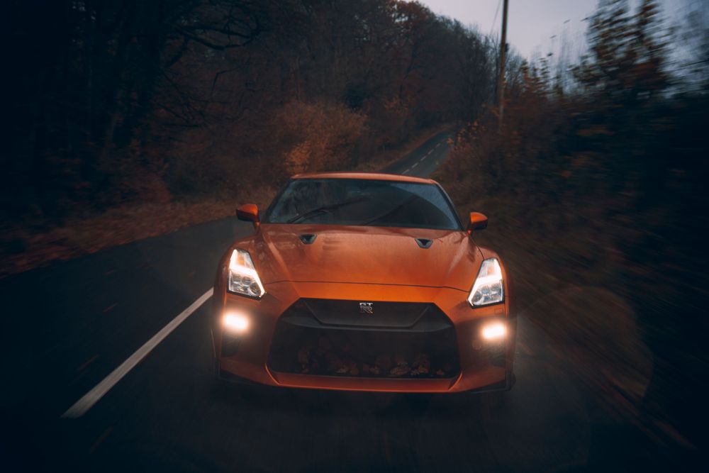 Orange sports car driving on dark forest road with glowing headlights illuminating the path ahead.