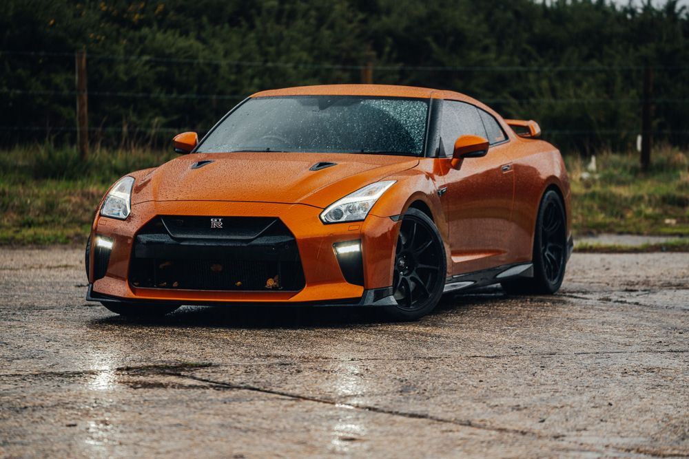 A bright orange Nissan GT-R sports car with black wheels parked on wet pavement with a forest backdrop.