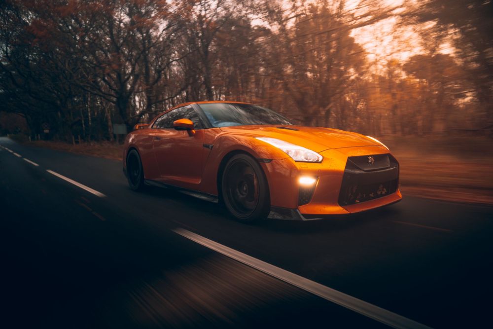 Orange sports car driving through misty forest road during golden sunset creating dramatic lighting effect.