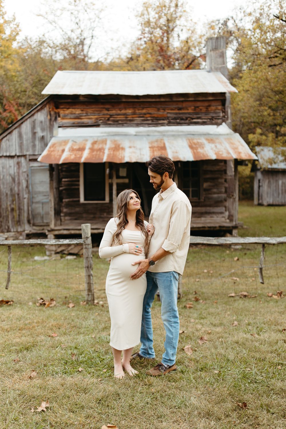 A couple poses in front of rustic wooden cabins during an autumn maternity photoshoot.