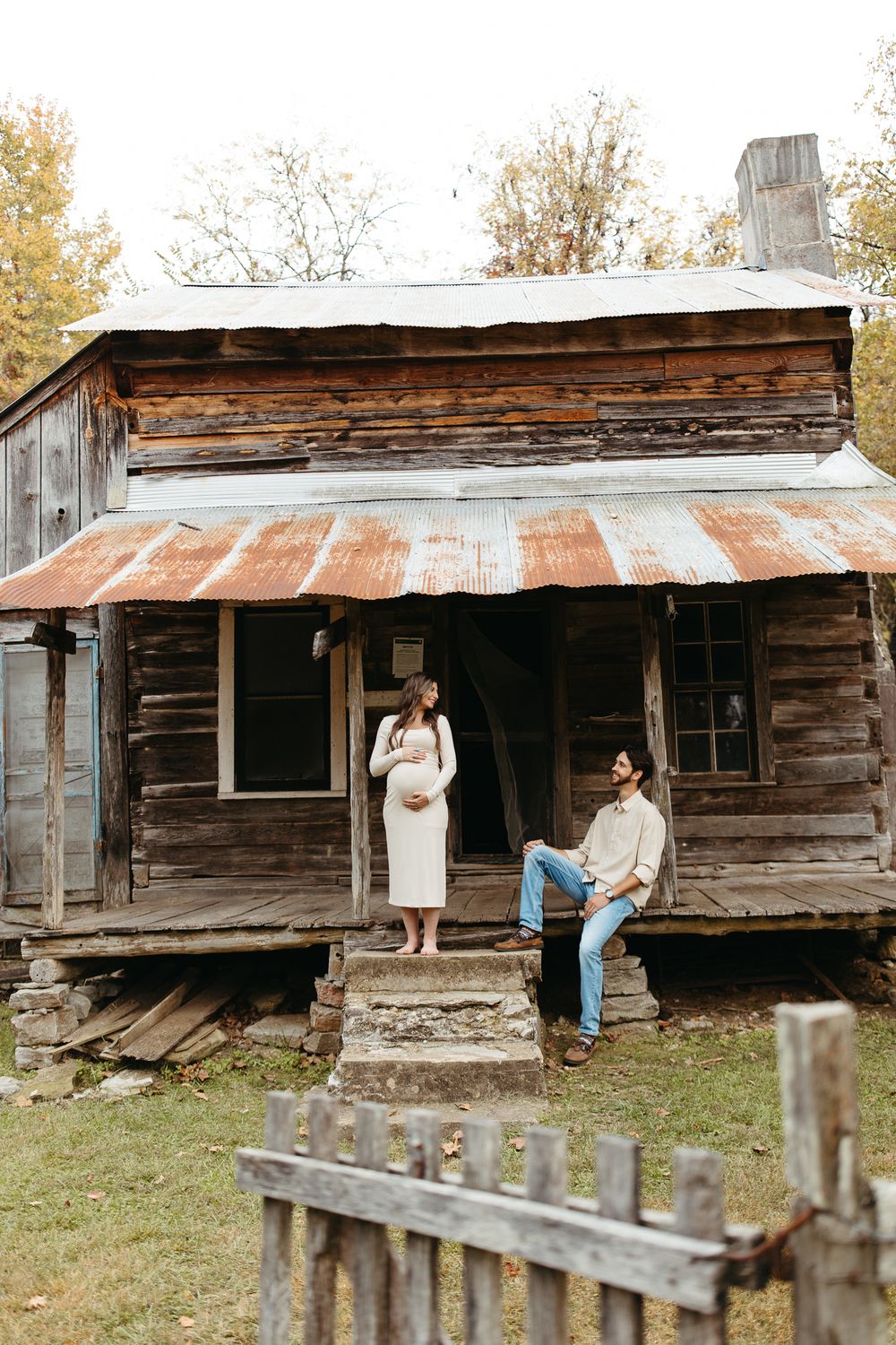 Rustic wooden cabin with weathered siding and tin roof sits in an autumn field surrounded by trees.