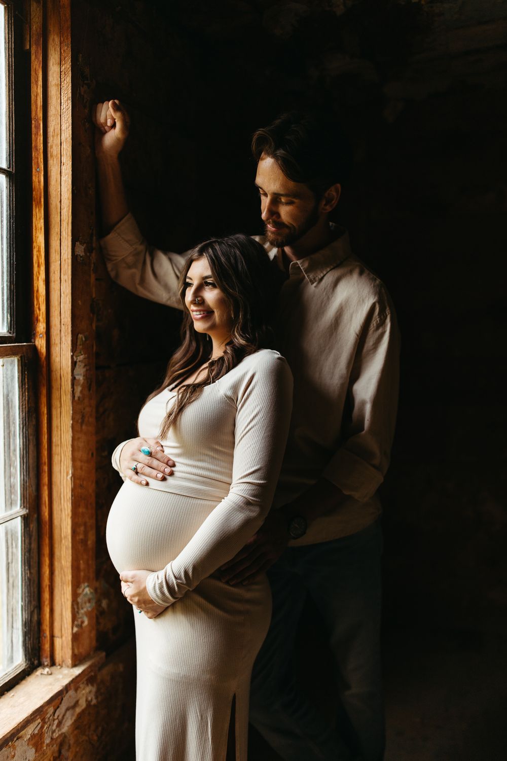 A couple stands by a window in a dark room, with the woman wearing a fitted white maternity dress in warm lighting.