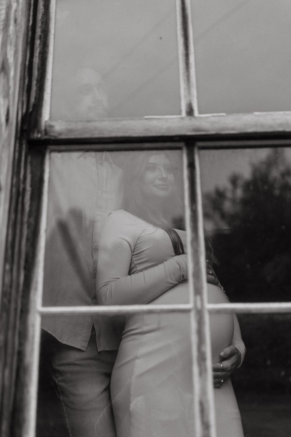 Artistic black and white maternity silhouette through window panes creating a dreamy, intimate atmosphere.