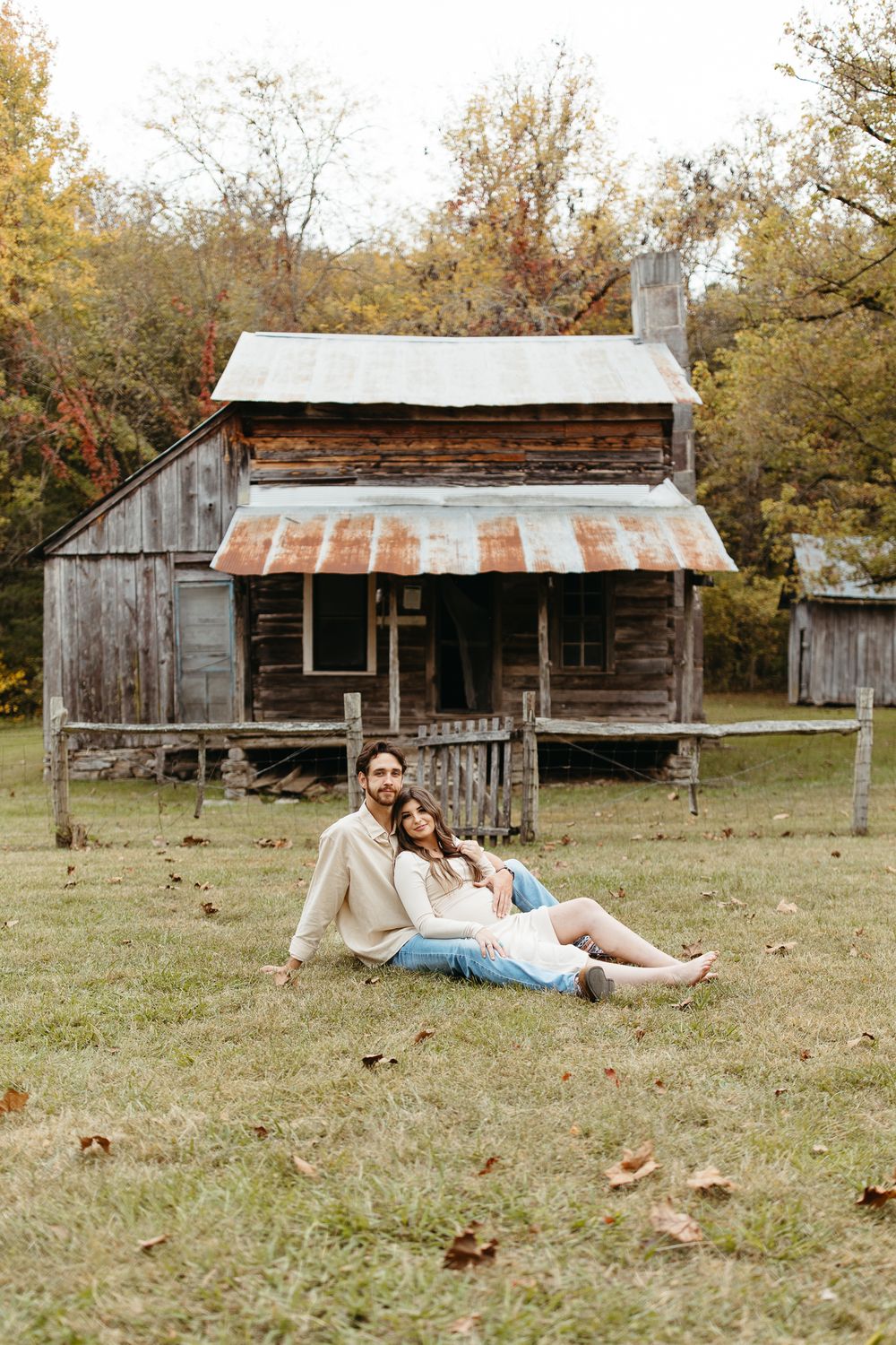 Couple relaxes on grassy lawn in front of vintage log cabin during fall season.