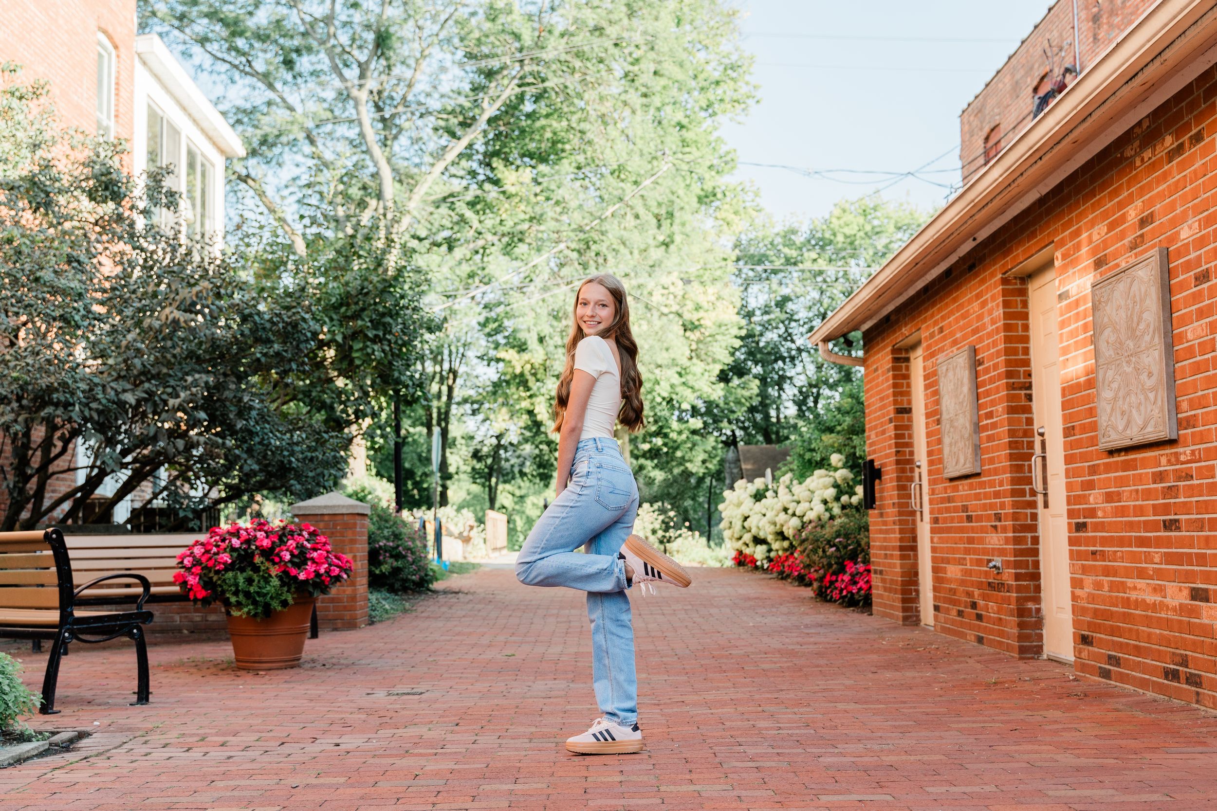Person walking down a brick pathway lined with red flower pots and trees on a sunny day.