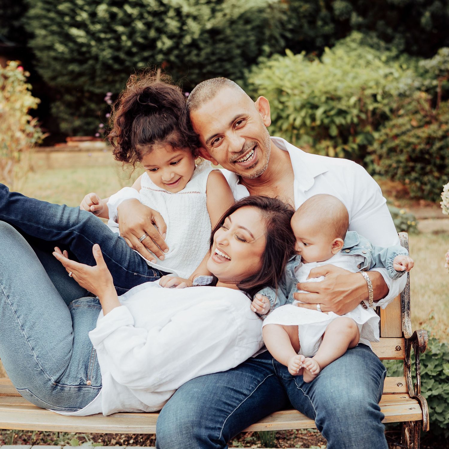 Family of 4 sitting outside on a bench laughing and cuddling.  The mum is lying down leaning on her husband & their children are around them
